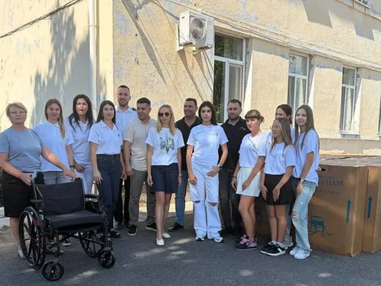 Група людей у білих футболках стоїть біля будівлі з візком і коробками крісел